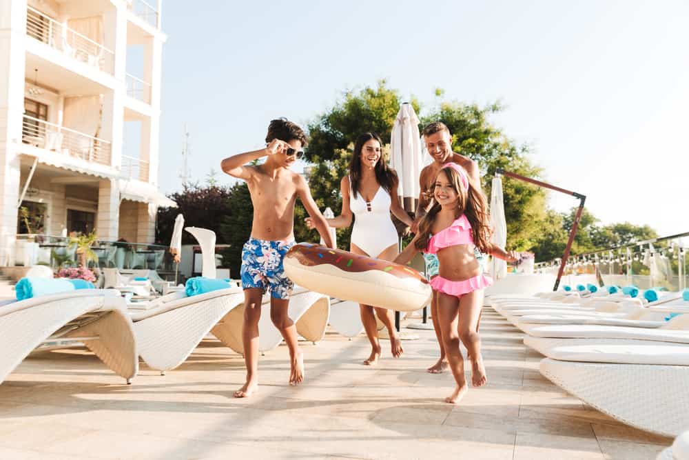 fun family vacation Image of happy caucasian family with children resting near luxury swimming pool with white fashion deckchairs and umbrellas outside hotel